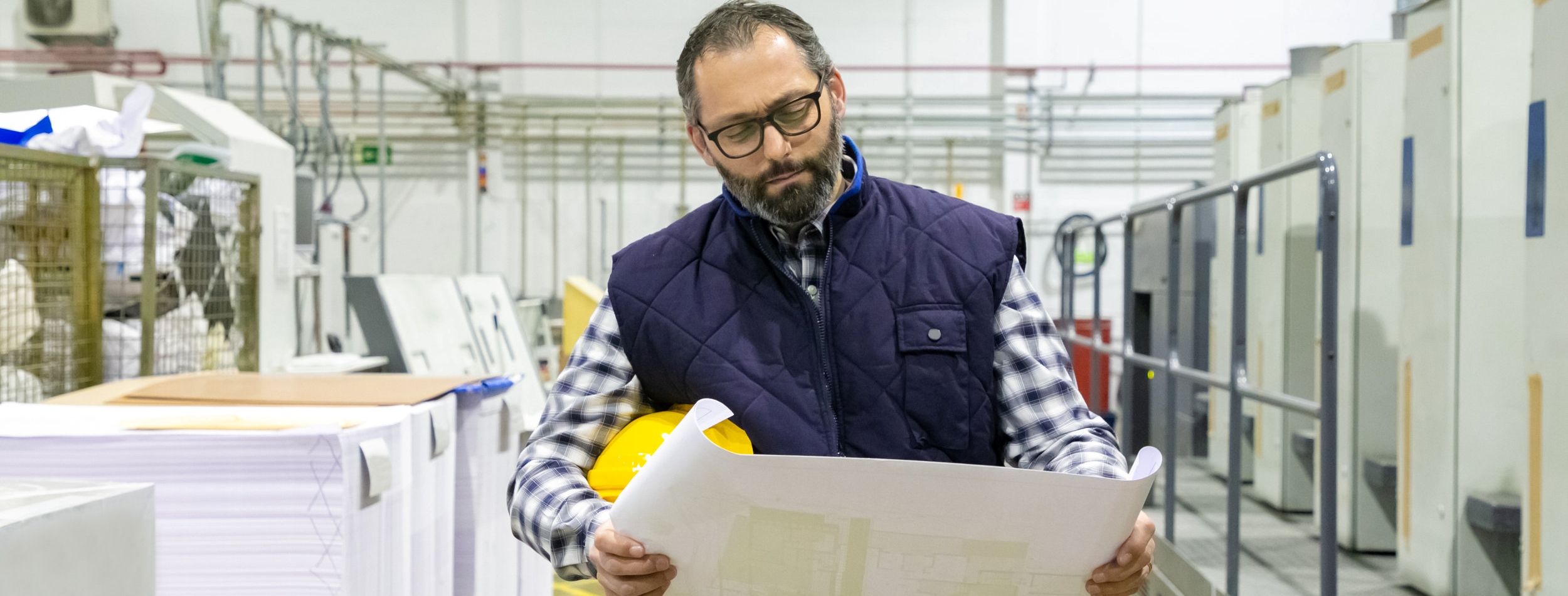 Front view of thoughtful technician inspecting blueprint. Professional printing worker with building plan at factory. Print manufacturing concept