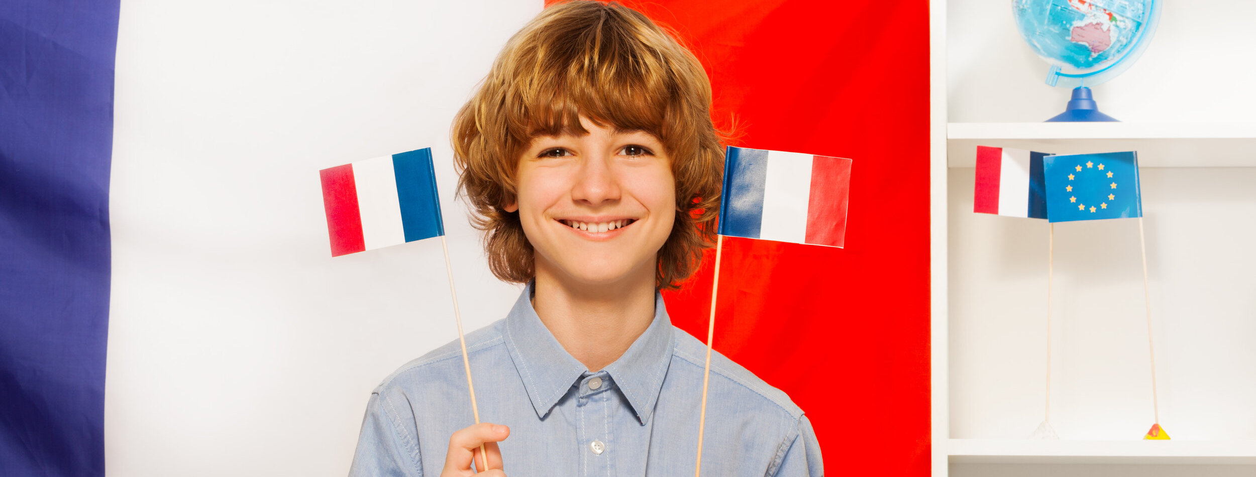 Smiling teenage boy, holding two flags in his hands, studying French language at the class